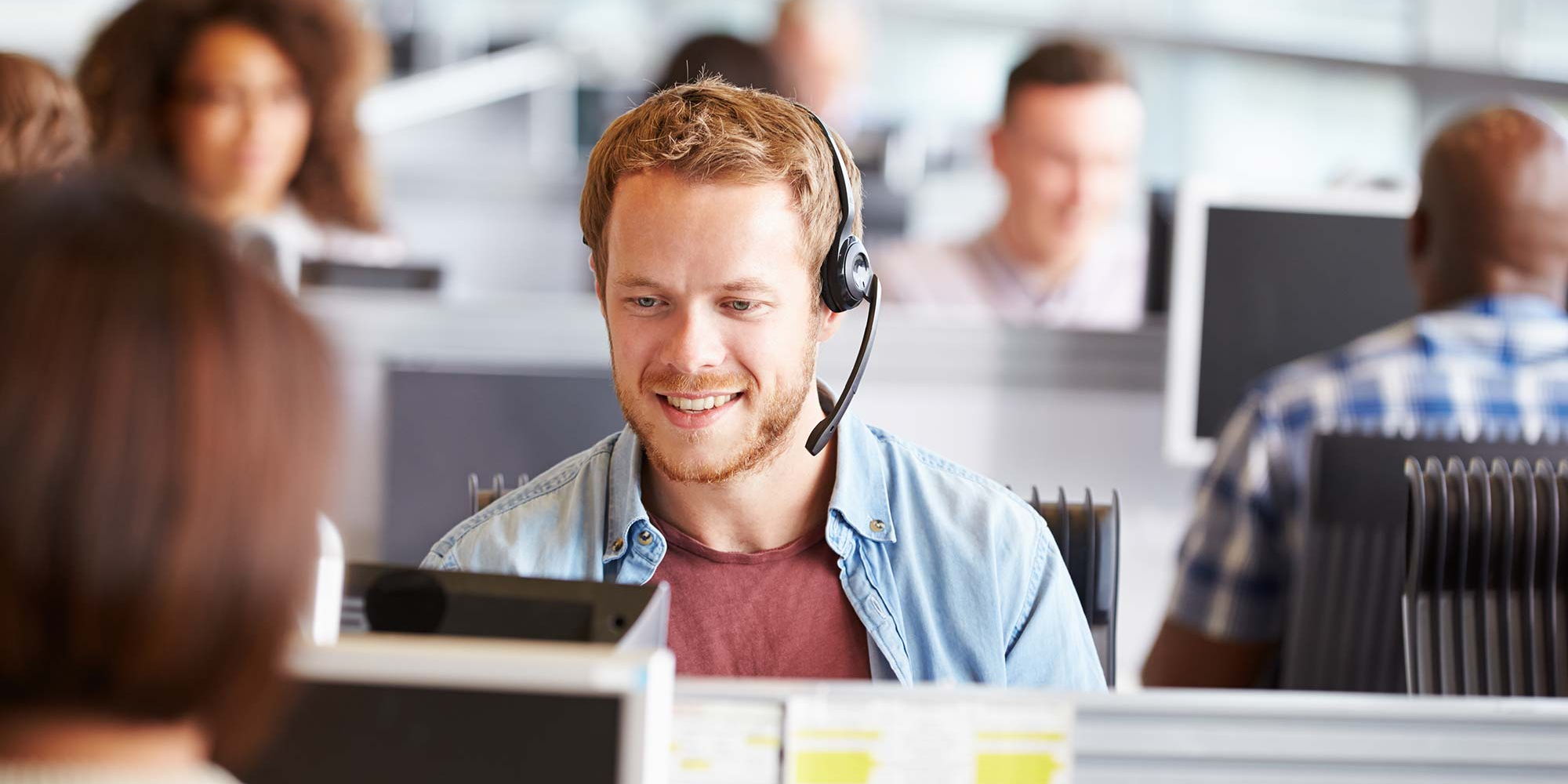 Ein freundlicher junger Mann mit Headset arbeitet im Support-Team, umgeben von Kollegen im Büro.